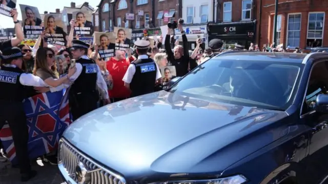 A parked car in front of a protest. People are being held back by police officers. They hold signs up of Starmer's face which say 'Keir Starmer Jew hater'