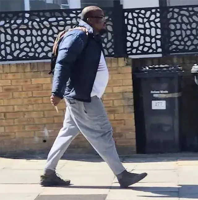 A screengrab of the suspect wearing a white t-shirt, dark jacket and grey pants walking down a street holding what appears to be a knife.