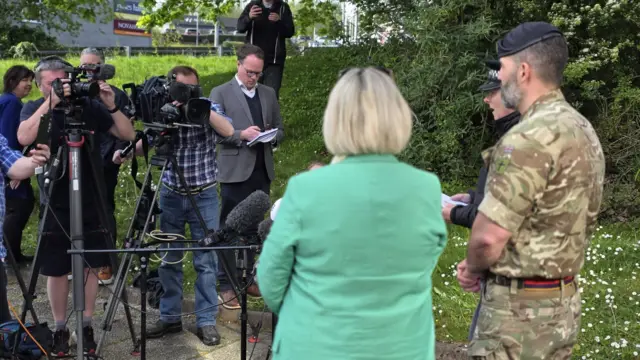The image shows an outdoor media briefing, with television cameras, microphones and journalists gathered on a grassy verge beside a path. In front of them, uniformed officials and a person in a green jacket appear to be speaking to the press, with trees and buildings visible in the background.