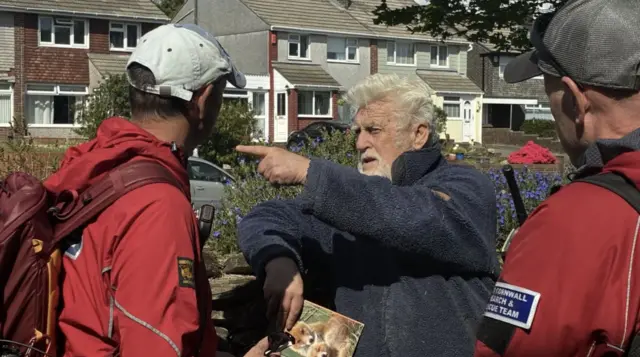 A bearded man wearing a blue top talks to two men in red unifoirms carrying rucksacks with houses behind them.