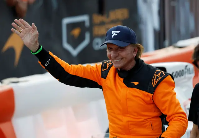 Emerson Fittipaldi, wearing an orange race uniform, waves at fans during the McLaren showrun event at Regatta Harbour in Coconut Grove in Miami