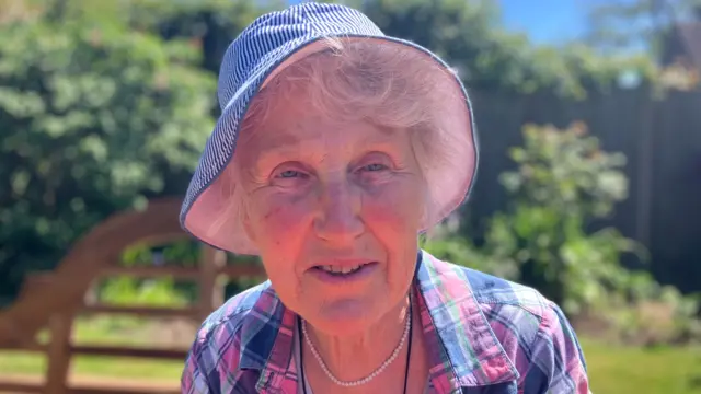 Woman is hat with pink and purple checked shirt with white necklace