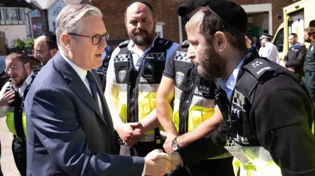 Starmer shakes hands with a Jewish Shomrim member who wears a security vest. Other Shomrim members look on