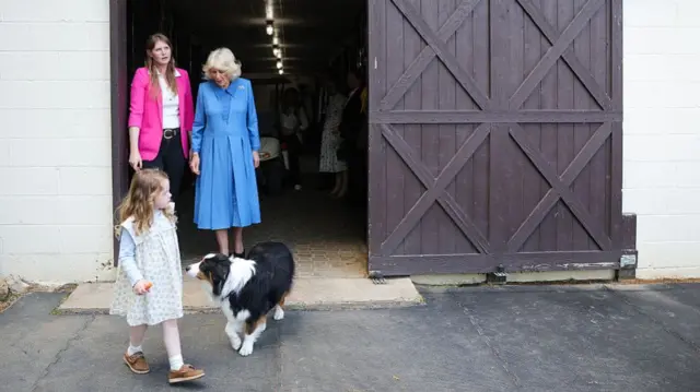 A woman in a bright pink jacket walks beside the Queen, dressed in blue, while in front of them are a little girl and a dog