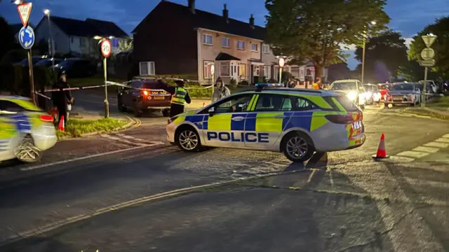 A police car in the middle of the road with a officer at the front speaking with a woman. There is a cordon in place nearby houses.