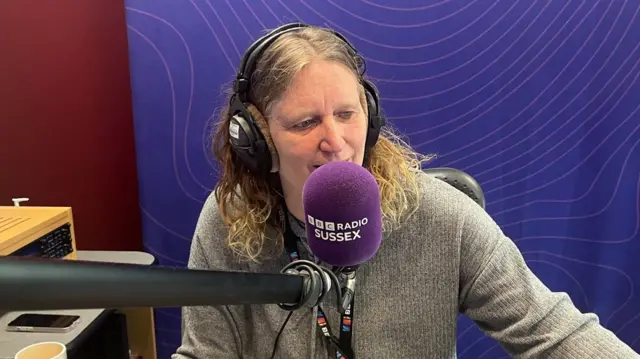 Sarah Gorrell looking to the right of the camera and sitting behind a purple BBC Radio Sussex microphone. She has brown, curly hair and is wearing black headphones. She is also wearing a grey jumper. She is sitting in front of a purple background with a swirly pattern.