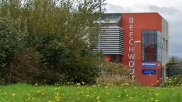 Beechwood Primary Academy - a red building with Beechwood on it in vertical lettering - behind green trees with grass in the foreground.