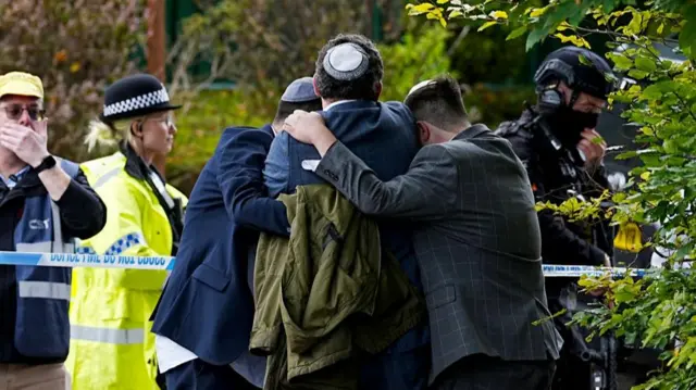Three men holding on to each other in an embrace, with backs to the camera. On the other side of police tape in front of them stands a uniformed officer, a security man and a police officer holding a gun