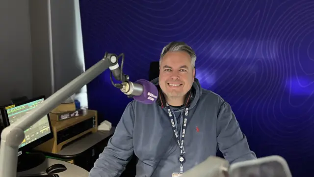 John Acres sitting at a radio desk. He is smiling at the camera withe a purple microphone in front of him and a purple backdrop behind.