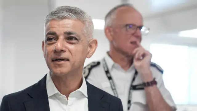 Sadiq Khan wears a black blazer and white shirt while Mark Rowley stands blurred in the background of a bright white room