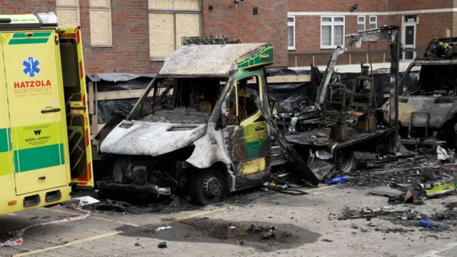 The wreck of a burnt out ambulance which sits in a car park, completely destroyed with debris around it. The back of a non burnt ambulance can be seen parked in front