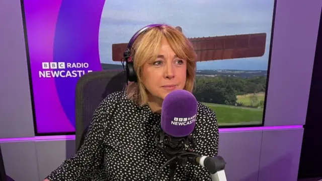 Anna Foster looking to the right of the camera with a slight smile. She has blonde, shoulder-length hair and is wearing black over-the-ear headphones and a black and white patterened shirt. She is sitting behind a purple microphone which reads BBC Radio Newcastle and is in front of a photo of the Angel of the North.