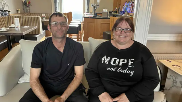 Two people in black tops one with a logo sit on a sofa with furniture around them.