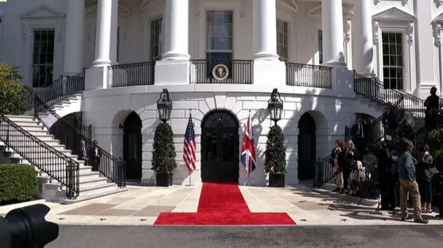 South Portico of White House with balcony and stairs leading down to an area with red carpet and US and UK flags, as reporters stand around