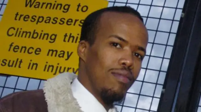 Image of Essa Suleiman, pictured in 2008, wearing a brown jacket with fur collar and white shirt and standing in front of a metal fence