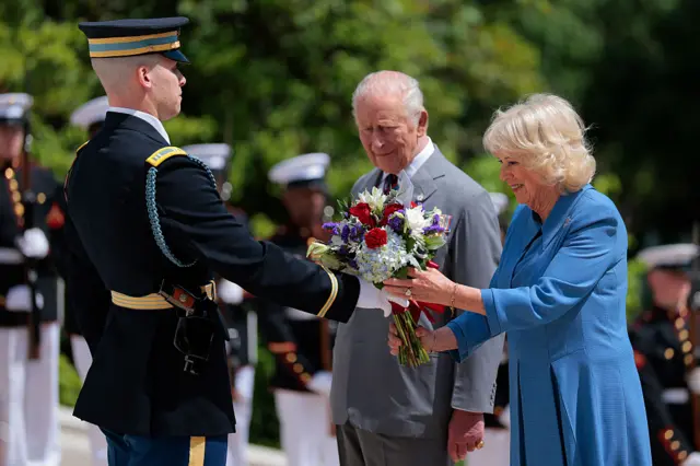 A military officer hands Queen Camilla a bouquet of flowers while King Charles looks on