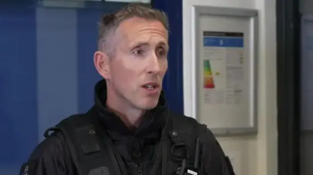 A police officer in police uniform. He is looking to the right. There is a blue and white wall behind him.