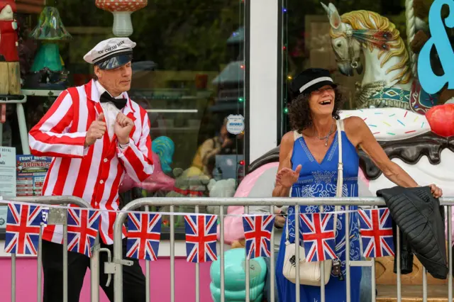 Two people wearing fancy outfits dance behind metal barriers. The barriers are draped with UK flags