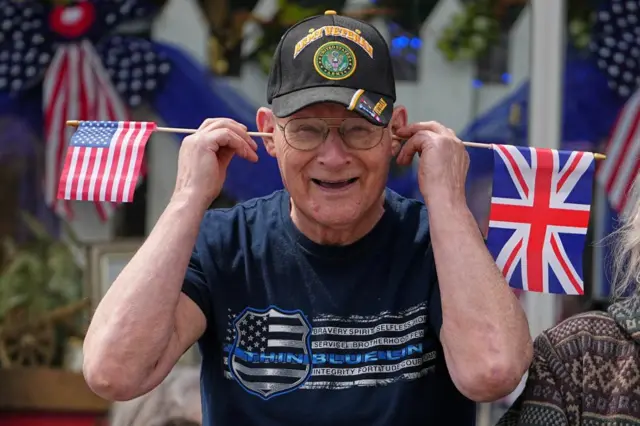 A man holds UK and US flags between his ears while waiting for Britain's King Charles and Queen Camilla attend a parade in Front Royal, Va