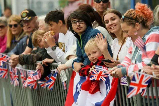 People stand behind metal barrier, covered in British flag bunting, as they attend a parade and block party