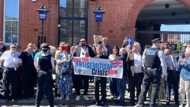 A crowd of protesters on a pavement lined up holding posters of Starmer's face and a big white banner that reads stop the antisemitism crisis now. There are two Metropolitan police officers standing in front of the crowd