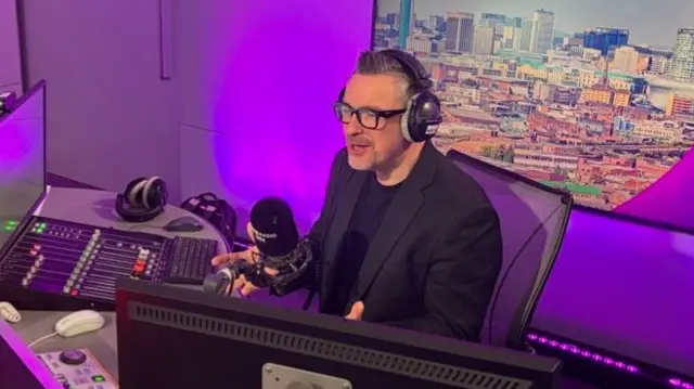 Ed James talking into a black micrphone while wearing black headphones. In front of him are a couple of computer screens and a soundboard. He has grey hair which has been gelled to stick up on the top and is wearing black glasses and a black blazer over a black t-shirt. There is a purple light behind him and a large photo of Birmingham's skyline.