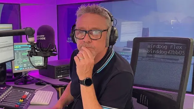 Tony Snell looking into the camera with his chin on his hand. He has grey, spikey hair and black glasses. He has a silver wedding ring on his hand, a smart watch and is wearing a blue polo shirt. He is sitting in an office chair behind a BBC Radio Merseyside purple microphone, two monitors and a soundboard.