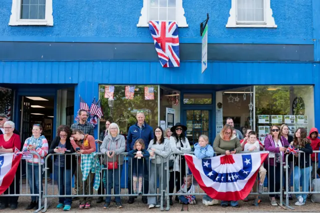 People line up the street as they wait for King Charles and Queen Camilla to appear - they're standing behind a metal barrier. A UK flag can be seen draped from the shop window behind them