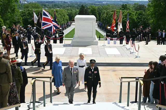 The royal couple ascending white steps with a dramatic vista of monuments, green lawns and military personnel with flags behind them