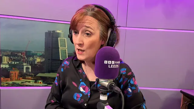 Gayle Lofthouse mid-speech looking to the left of the camera. There is a BBC Radio Leeds purple microphone in front of her and she is wearing black over-the-ear headphones. She is wearing a black shirt with blue and purple flowers on it and has brown hair which falls just below her chin. Behind her is a photo of the Leeds skyline.