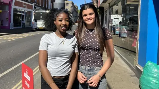 Salma Osman and Alicia Hassan stood next to each other on a high street. Salma is on the left and is wearing a grey t-shirt with black jeans. Alicia is on the right and is wearing a purple top with white polka dots on it and blue jeans. Both women have sunglasses on top of their head and are smiling for the camera.