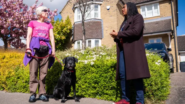 Kemi Badenoch talking to a woman on a residential street. The woman is wearing a pink t-shirt and has a black dog on a lead. Behind them are a low hedge and a brick house