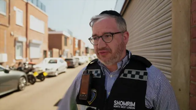 Headshot of Isaac who wears a Shomrim vest and stands in front of a street where there are parked cars and garage doors