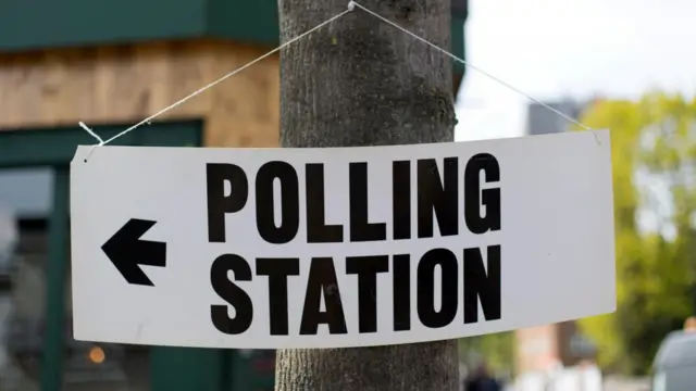 A close-up of a white sign with a black arrow pointing left and black writing in capital letters reading: POLLING STATION. It is hanging from string tied through holes at the top two corners, with the string looped round a nail in a tree trunk. The background is blurred