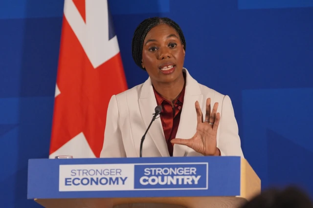 Kemi Badenoch speaking behind a podium which has a blue front and reads 'Stronger Economy Stronger Country'. She is gesitucualting with her hand and lookoing to the right. She is wearing a cream blazer and sily red shirt. Her dark, braided hair is tied behind her head. She is standing in front of a Union Jack and blue background.