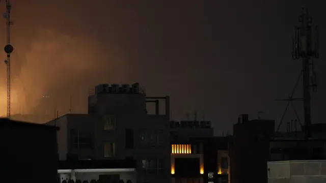 Smoke rises in a night sky over blackened buildings with a few lights on inside.
