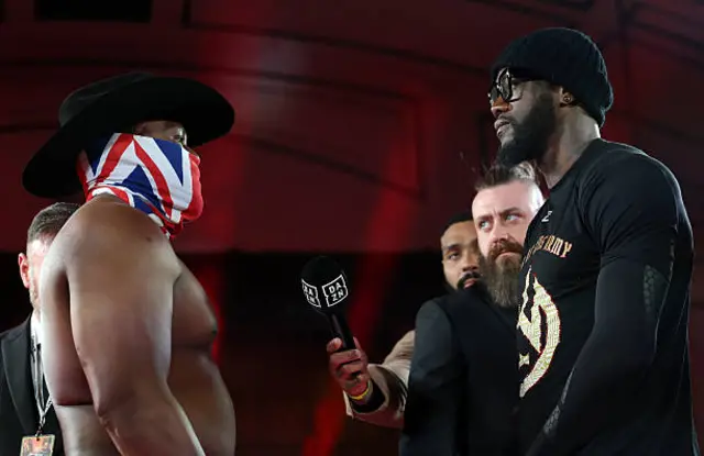 Derek Chisora has a Union Jack covering over his face and is shirtless as he stares at Deontay Wilder who is wearing a black t-shirt, glasses and hat