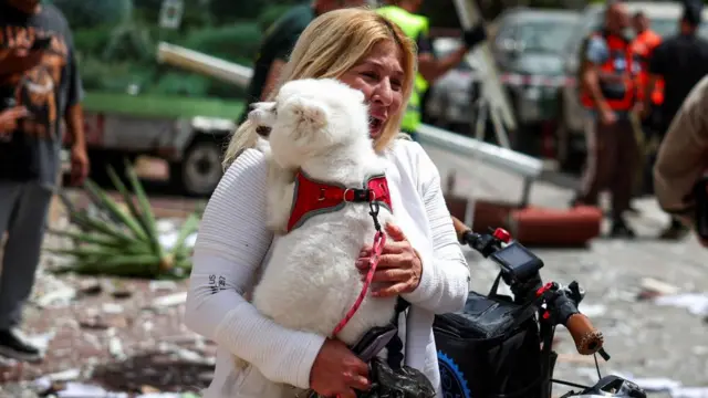 एक महिला जो व्याकुल दिखाई दे रही है वह मलबे और आपातकालीन कर्मचारियों के बीच एक छोटे सफेद कुत्ते को ले जा रही है