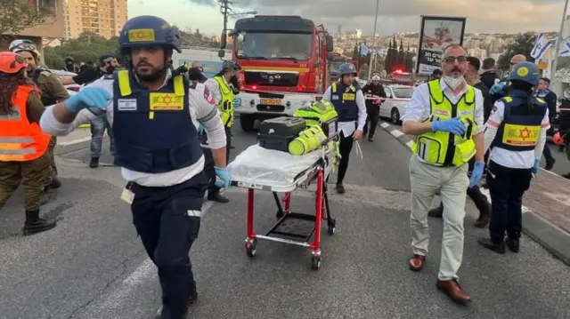 Israeli emergency responders wheel a stretcher down a city road as police, a fire truck, and bystanders surround the scene.