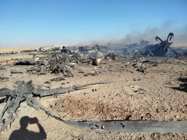 A burnt wreck pictured in a bare expanse of land. The strewn debris is charred and smoke is rising from the ground. It is daytime beneath a clear blue sky. The shadow of the photographer can be seen in the bottom left of the image.