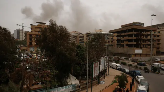 Smoke rising above a row of buildings, some blown-out, in Beirut. On the street people look up at the smoke behind a row of trees.