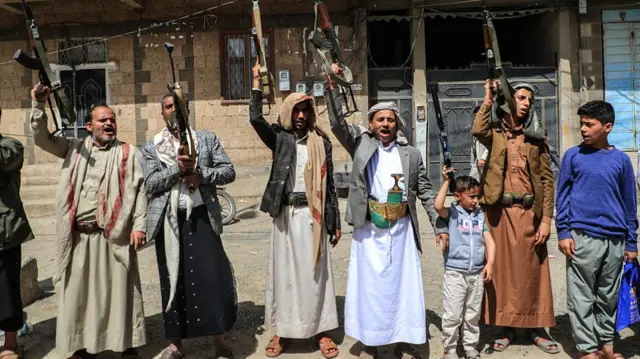 a line of men and boys, wearing long tunics and sandals, holding up rifles on the side of the street