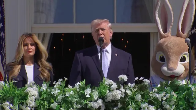 Donald Trump speaking into microphone. To his left is Melania Trump, to his right a large easter bunny in costume