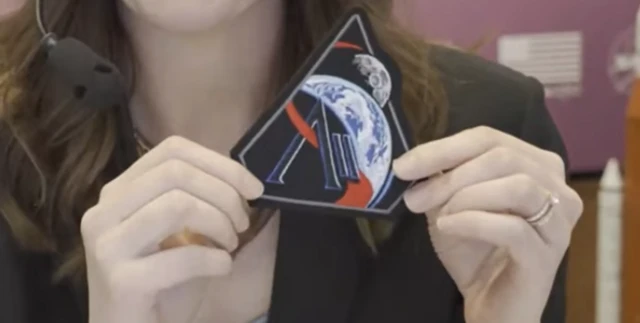 A triangular patch showing stylised representations of the Earth in front of the Moon, with the AII logo of Nasa's Artemis II mission is held by a person.