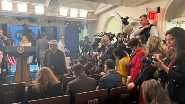 Inside press room of White House. Some reporters are sitting down on wooden chairs while the stage set up concludes, others stand with mics and cameras