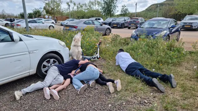 several adults and kids taking cover on the ground next to a vehicle as their dog howls beside them
