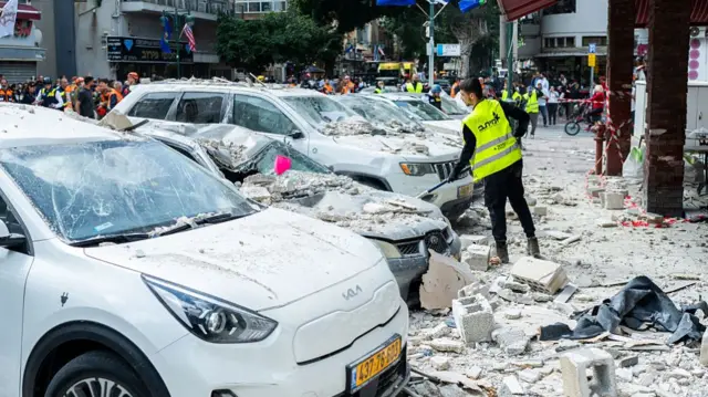 a man in a green safety vest looks at damaged cars covered in rubble on the side of a city street, with a crowd in the distance