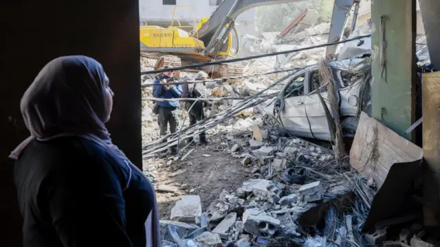 A woman watches as a bulldozer clears the rubble of destroyed buildings following an Israeli trike near the Rafik Hariri University Hospital (RHUH), in the Jnah District of Beirut, Lebanon.