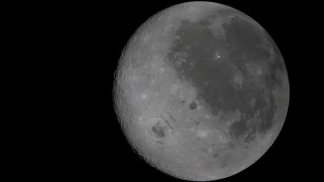 A fully illuminated view of the Moon where the near side can be seen on the right by the dark splotches. The background is pitch black and there are craters visible on the surface of the moon