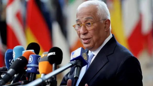 António Costa is shown speaking behind a bank of microphones. He is wearing a dark blue suit with a white shirt and light blue tie.